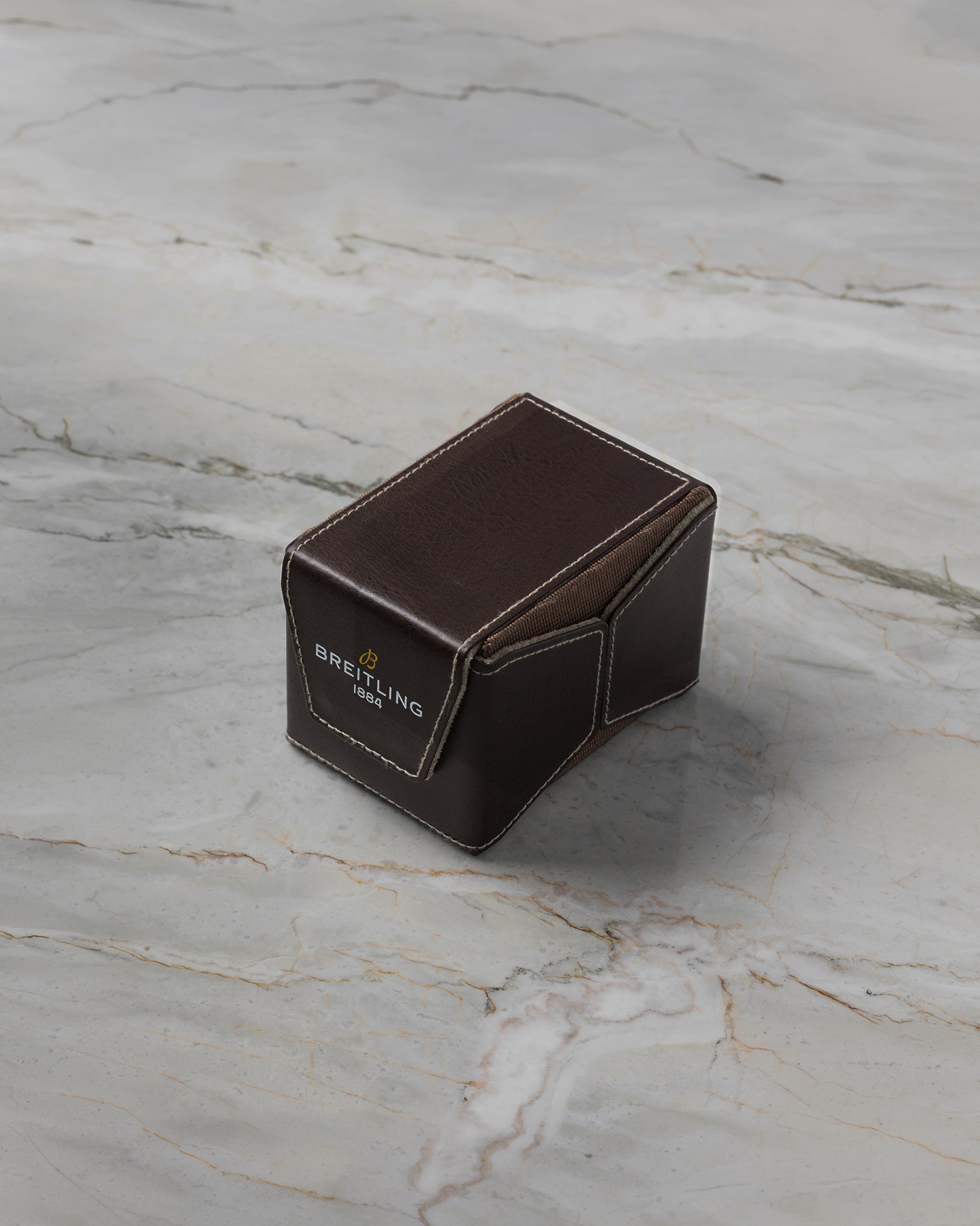 A three-quarter high-angle shot of the official folding dark brown textured leather watch travel case resting closed on a light grey marble surface