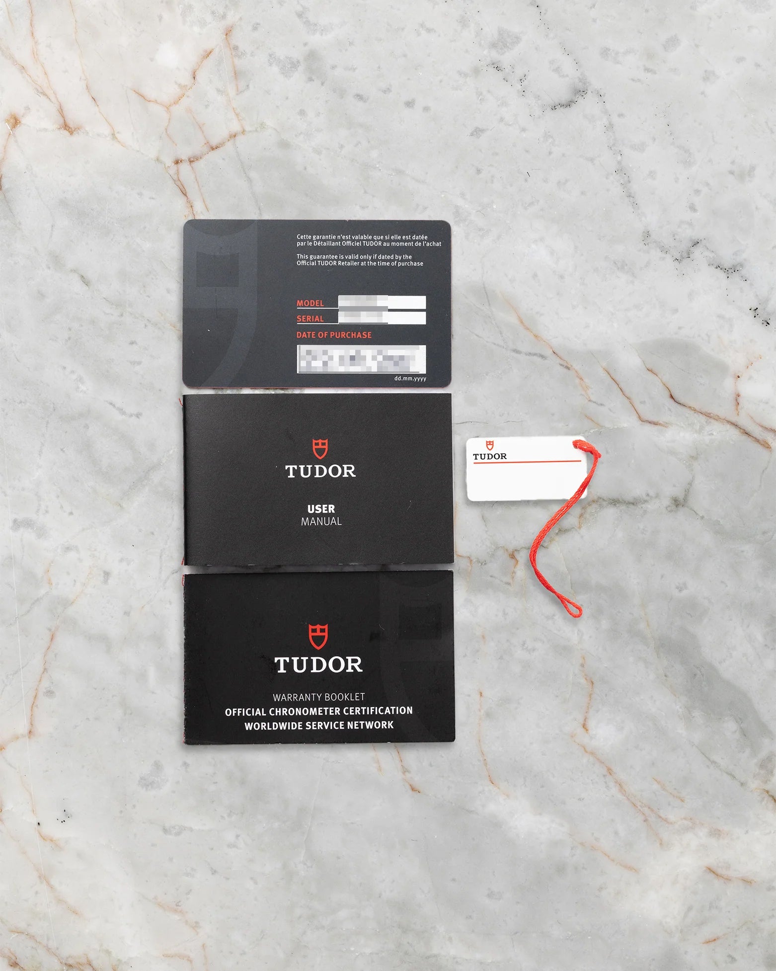 Official Tudor international warranty documentation cards resting stacked next to a white plastic retail hang tag on a marble surface
