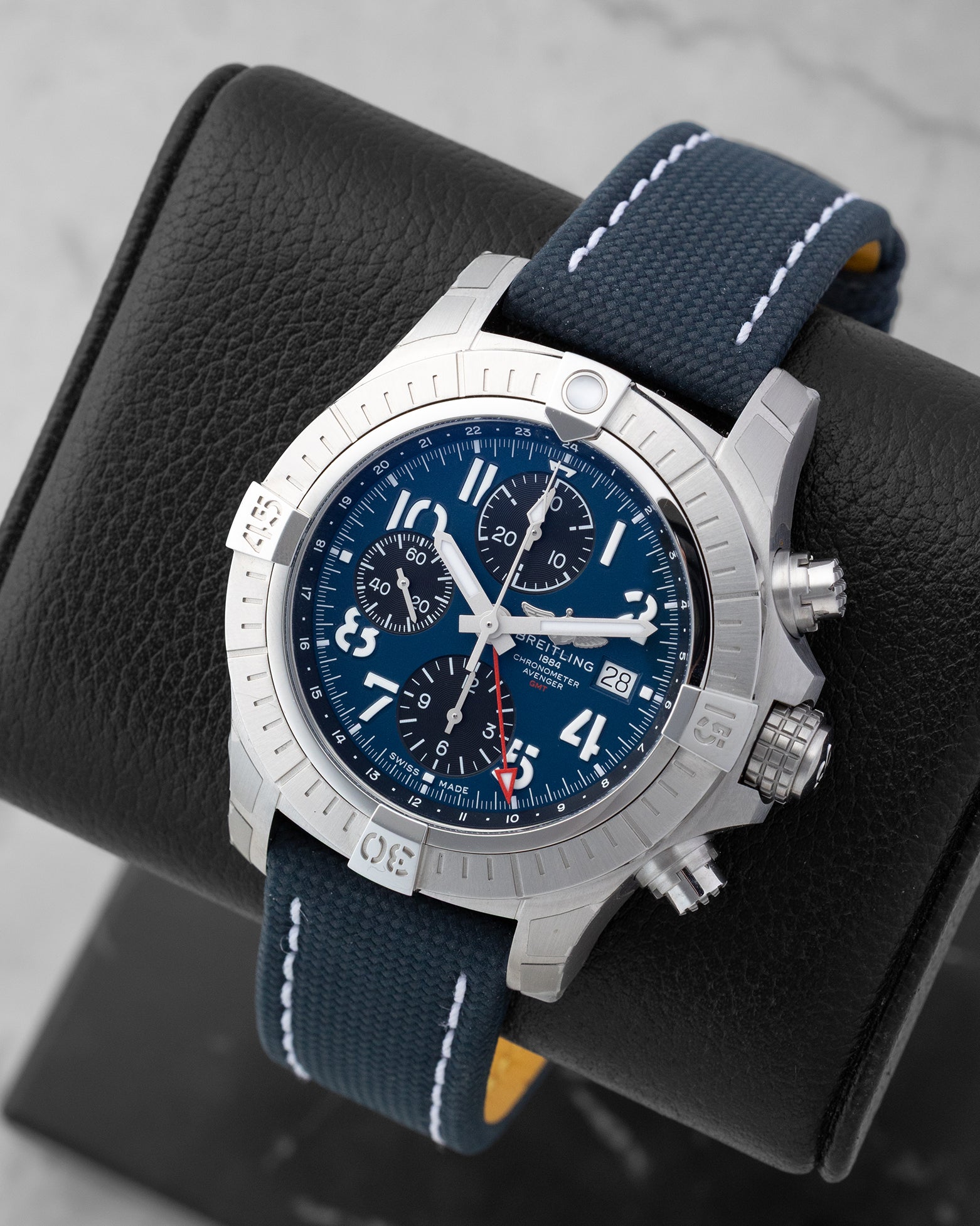 Angled front view of a preowned Breitling Avenger Chronograph GMT A24315101C1X1 featuring a blue stencil dial, red GMT hand, and blue leather strap with white stitching