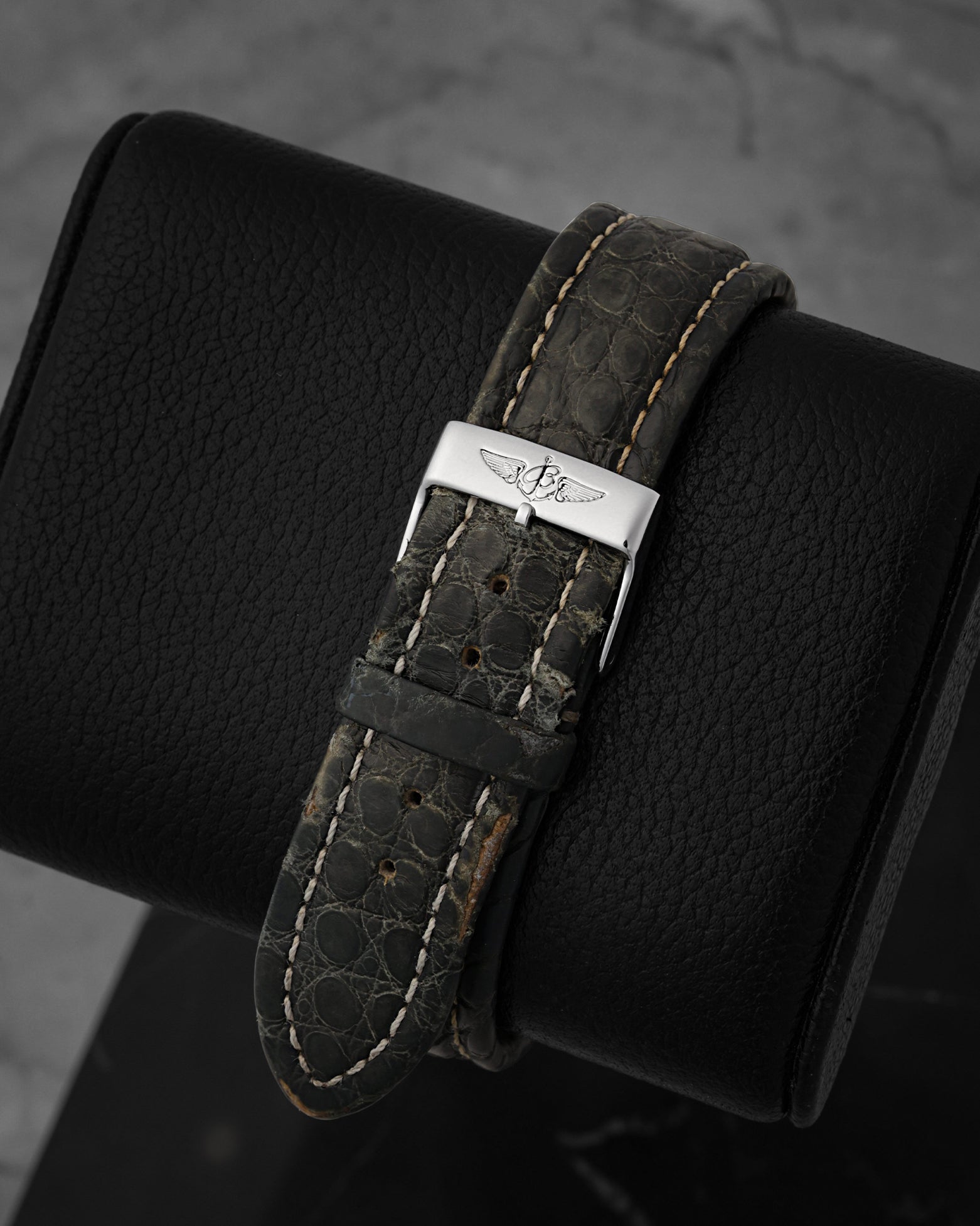 Close-up view of the premium dark grey textured exotic leather strap on a watch cushion, focusing on the polished stainless steel pin buckle clasp engraved with the classic Breitling script logo