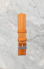 Included secondary vibrant orange textured leather replacement watch strap with a steel tang buckle laid out on a white marble backdrop