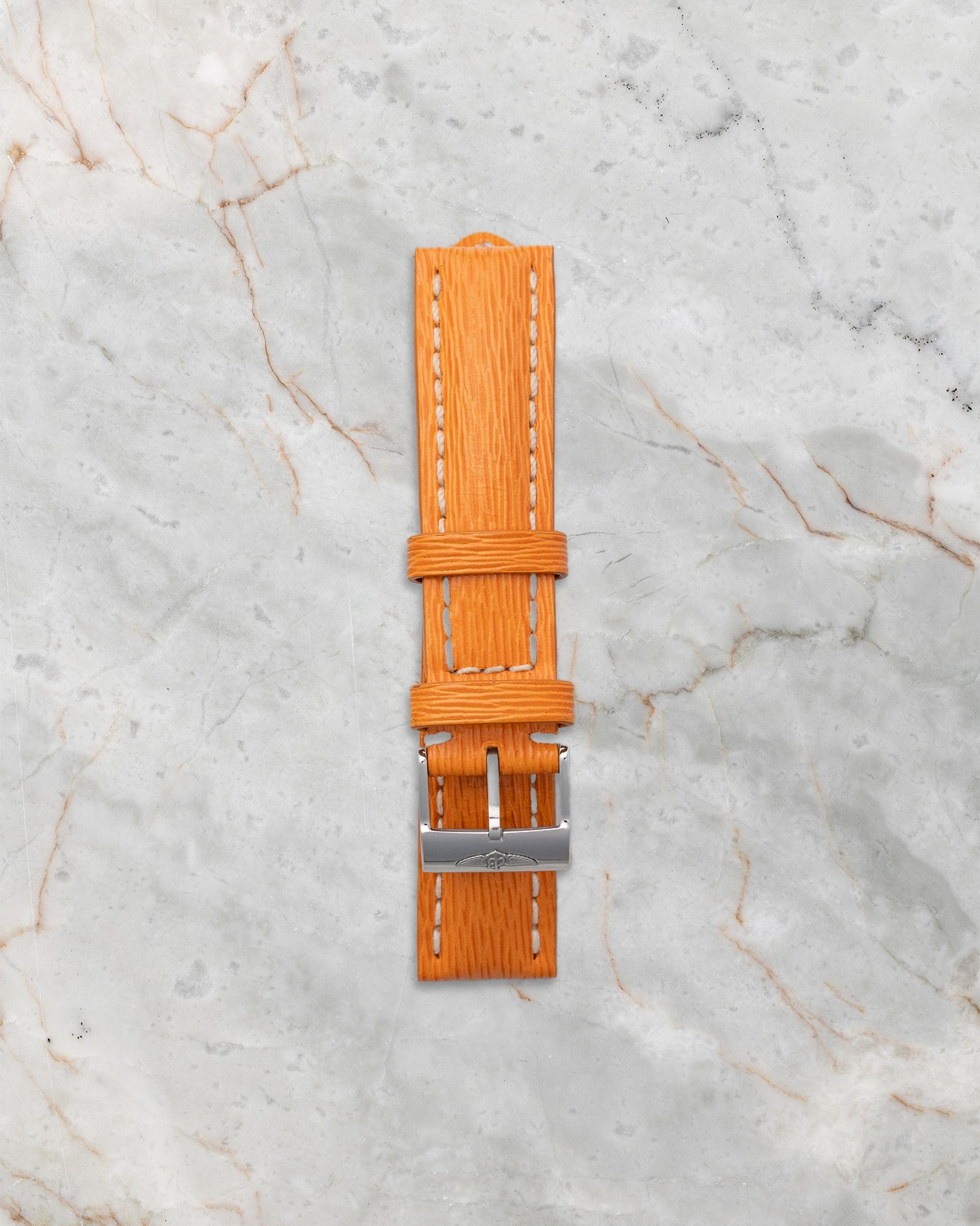 Included secondary vibrant orange textured leather replacement watch strap with a steel tang buckle laid out on a white marble backdrop