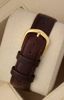 A rear view of the smooth mahogany brown leather watch strap focusing on the classic high-polished yellow gold-plated pin buckle clasp resting on a beige display cushion