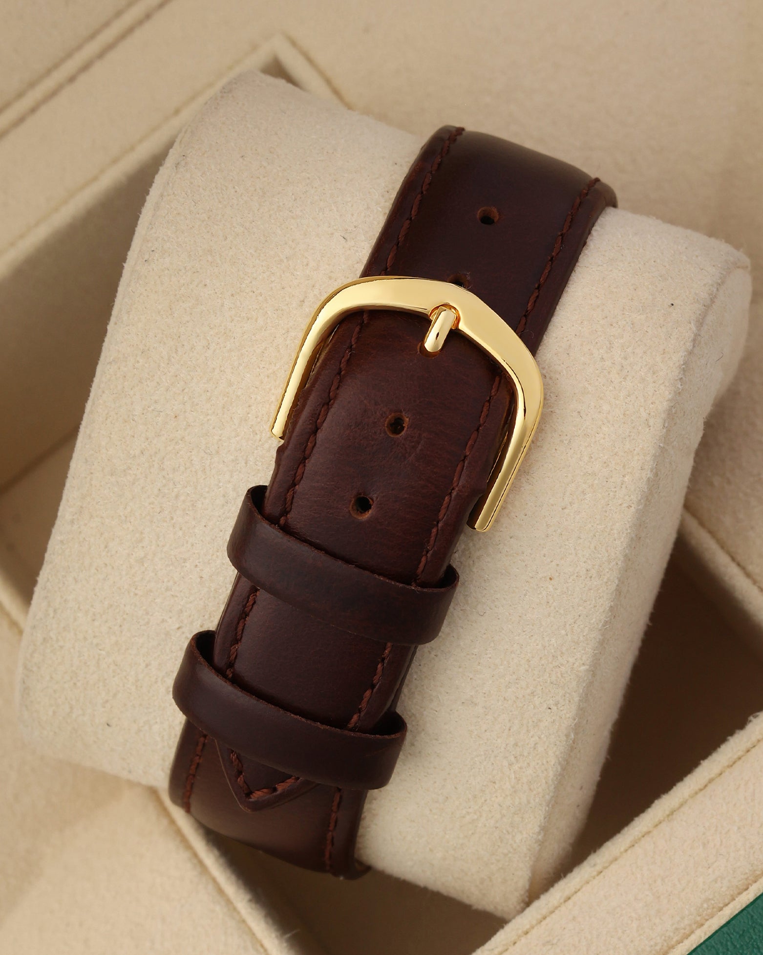 A rear view of the smooth mahogany brown leather watch strap focusing on the classic high-polished yellow gold-plated pin buckle clasp resting on a beige display cushion