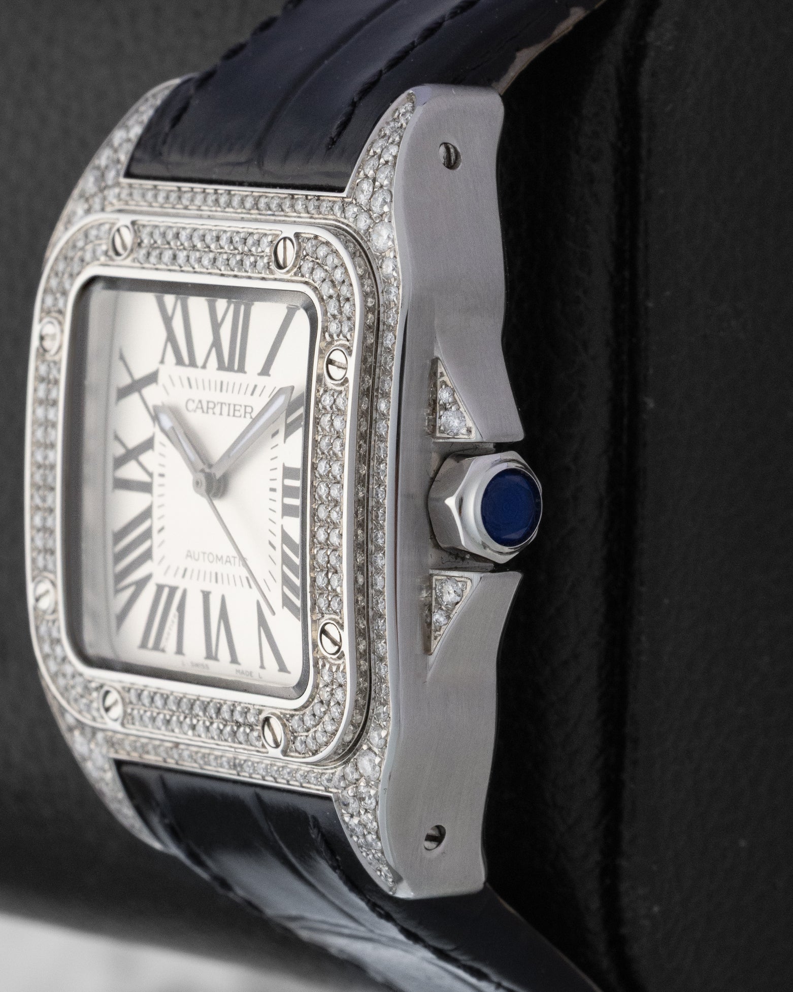 Right side profile of a diamond-encrusted Cartier Santos watch highlighting the faceted crown set with a blue synthetic spinel