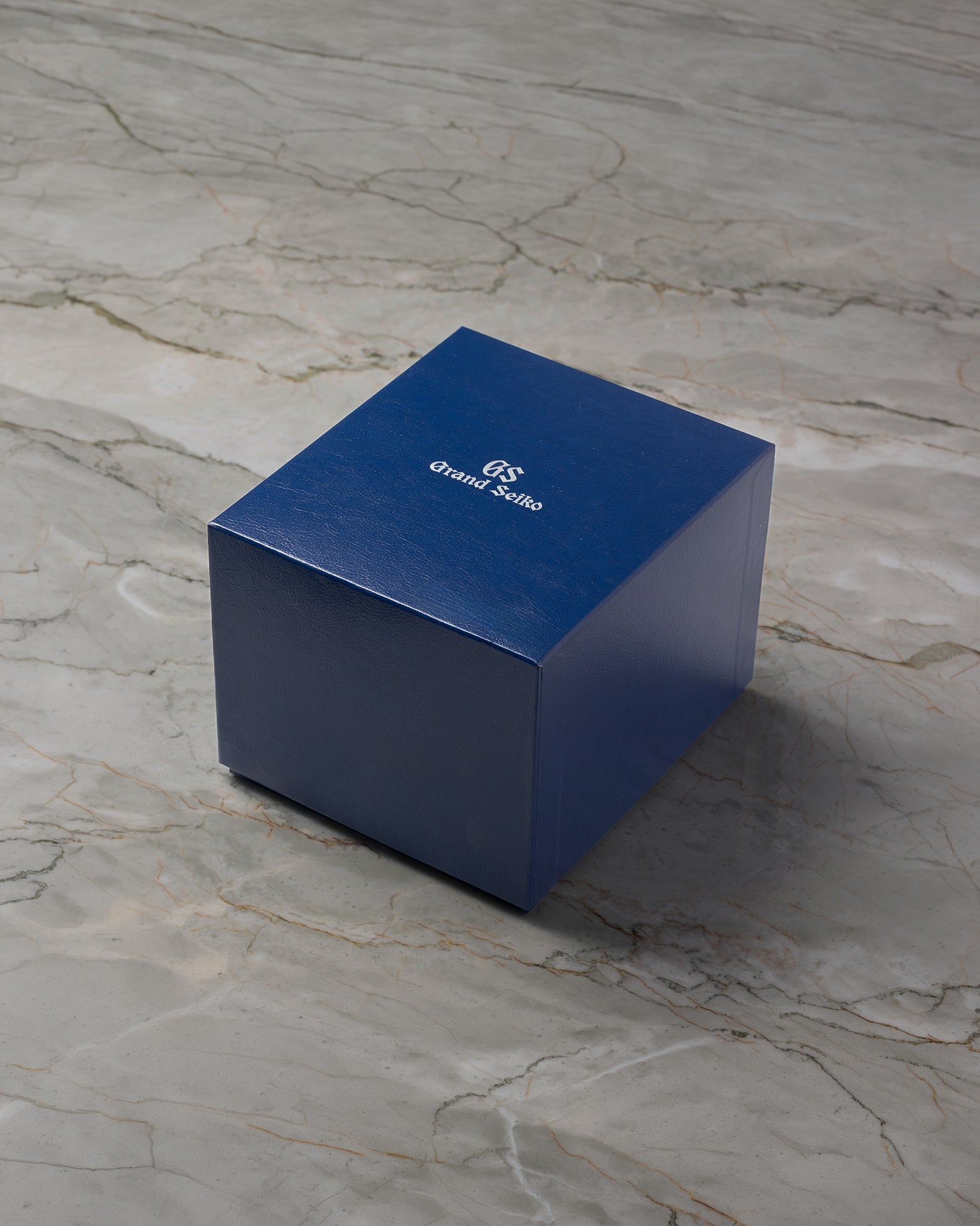 A high-angle three-quarter shot of the official minimalist matte navy blue Grand Seiko watch box resting closed over a light grey marble surface