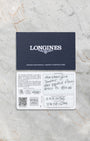 "The official blue and white Longines international warranty card with handwritten dealer purchase details laid flat on a marble background