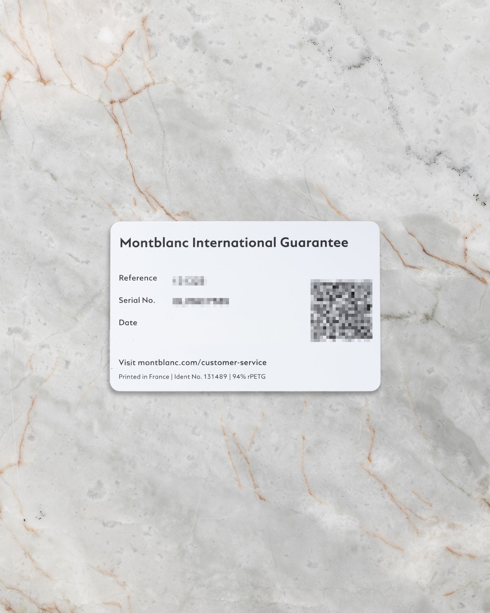 A top-down flat lay view of the official white Montblanc international guarantee card showing the printed reference block and a scannable QR code on a grey marble surface