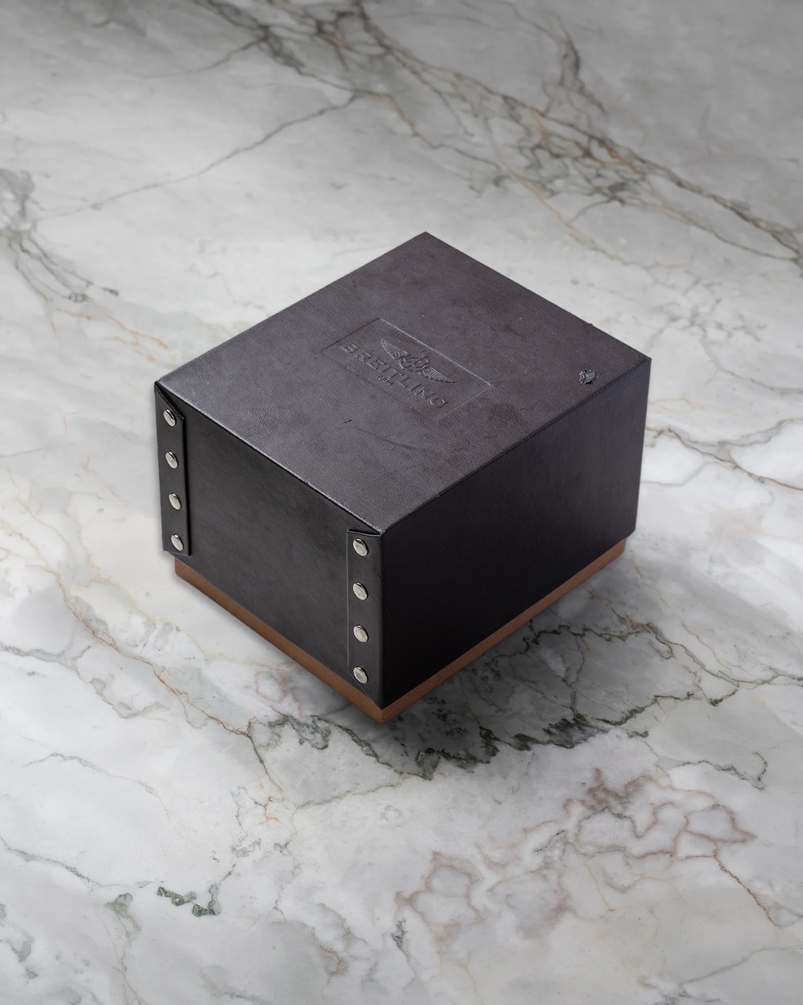 Original square textured dark grey Breitling luxury watch presentation box with stud details displayed on a premium white marble surface