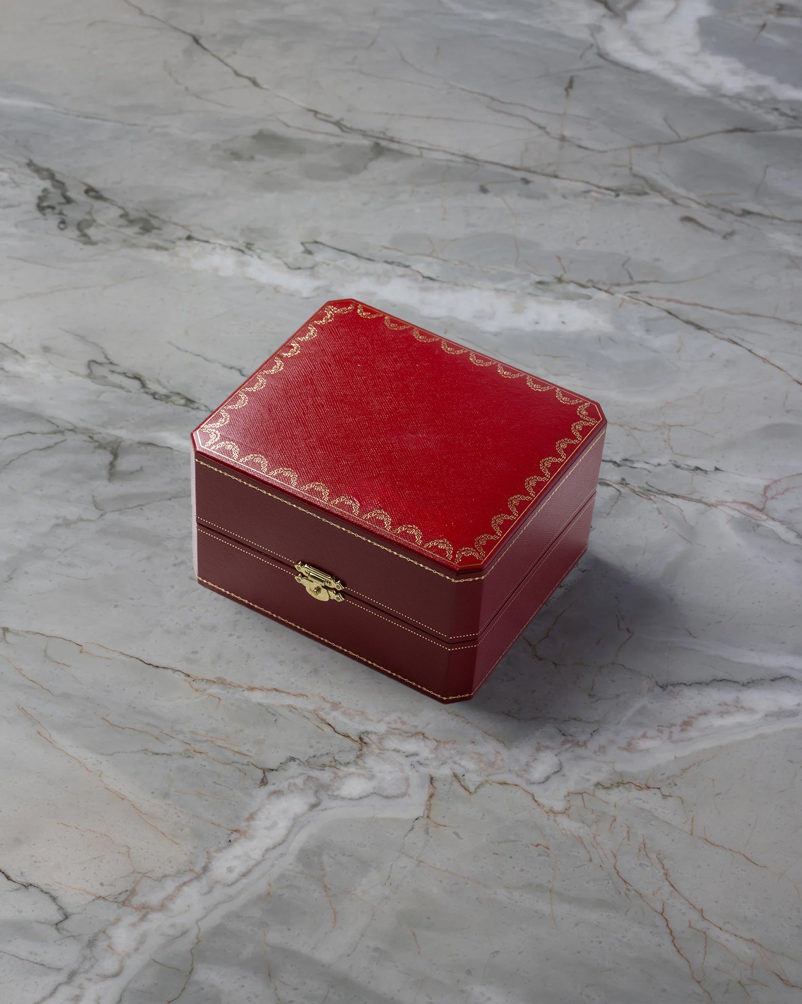 Original square textured red leatherette Cartier luxury watch presentation box featuring gold filigree detailing, displayed on a white marble surface