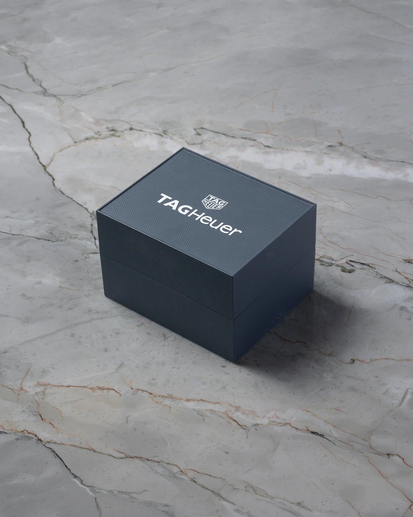 Original square matte grey-blue textured TAG Heuer luxury watch presentation box displayed on a premium white marble backdrop