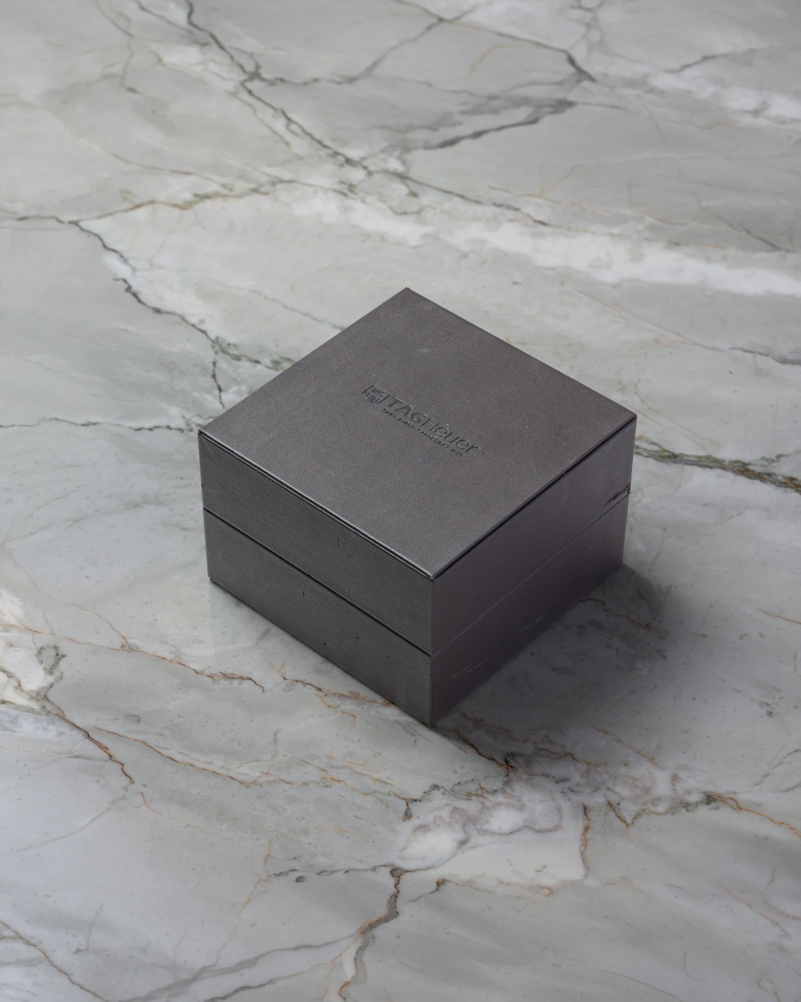 Original square textured brown TAG Heuer luxury watch presentation box displayed on a premium white marble surface