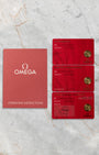 A top-down flat lay view of the official red Omega user operating manual alongside the triple red cards including the international warranty, pictogram, and master chronometer certification