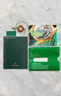 A flat lay view of the original 2002 era printed Rolex registration warranty certificate paperwork, green wallet sleeve, and green chronometer hangtag on a marble surface
