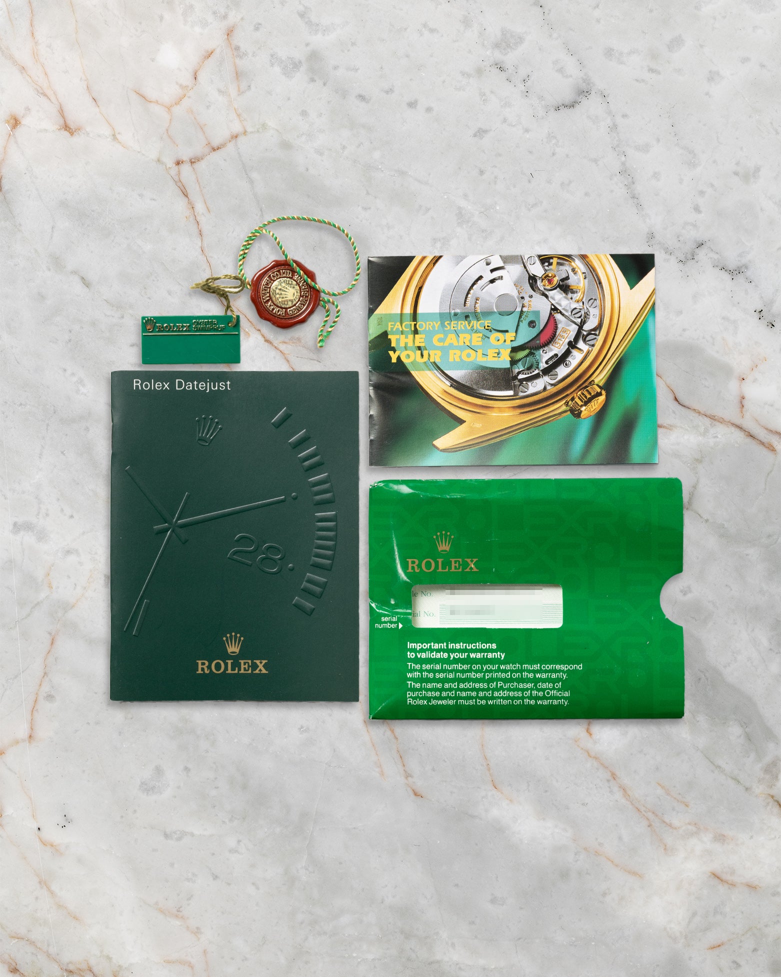 A flat lay view of the original 2002 era printed Rolex registration warranty certificate paperwork, green wallet sleeve, and green chronometer hangtag on a marble surface