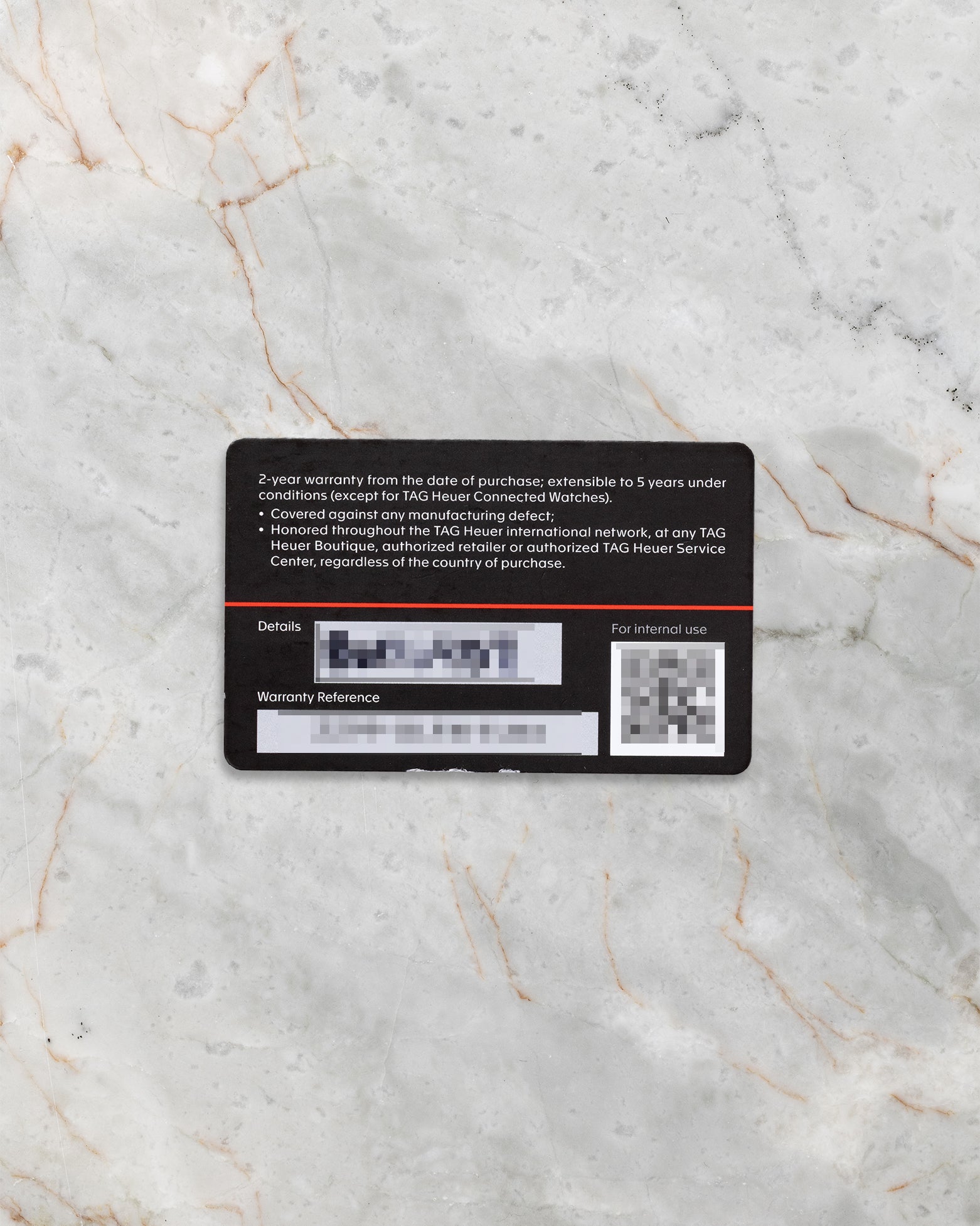 The back of the official black TAG Heuer international warranty card showing QR code and printed serial numbers flat on a marble surface