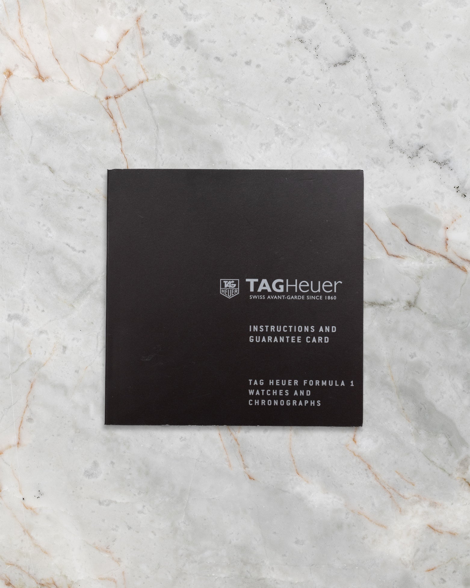 The official black TAG Heuer instructions and guarantee booklet laid flat centered on a white marble background