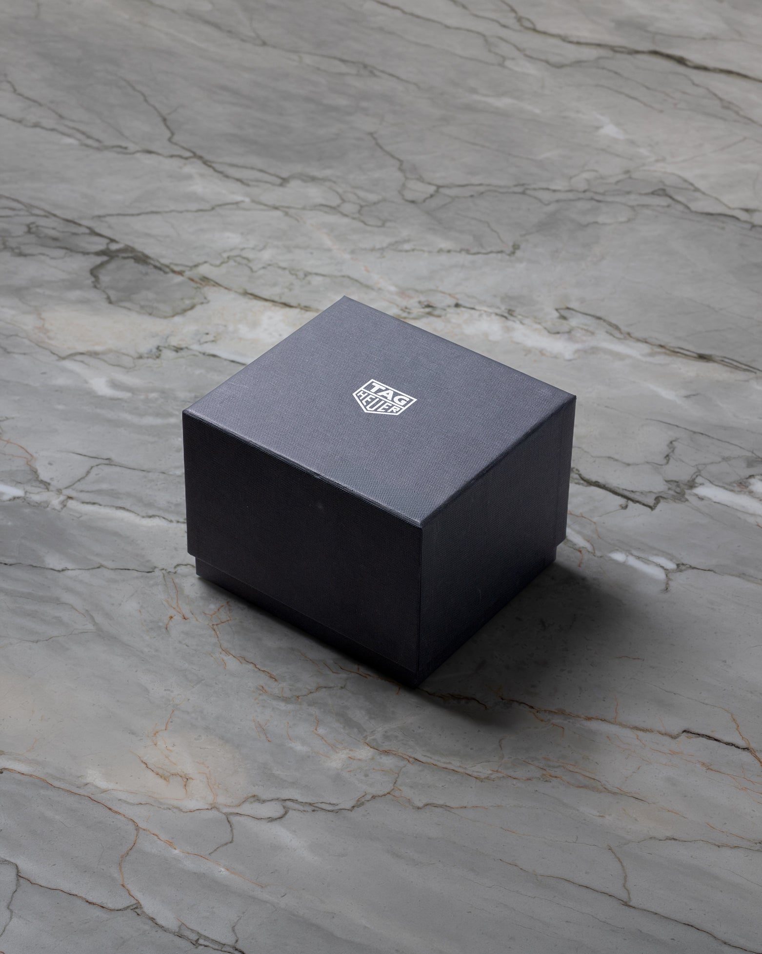 A closed square matte black TAG Heuer luxury watch presentation box sitting centered on a grey marble background