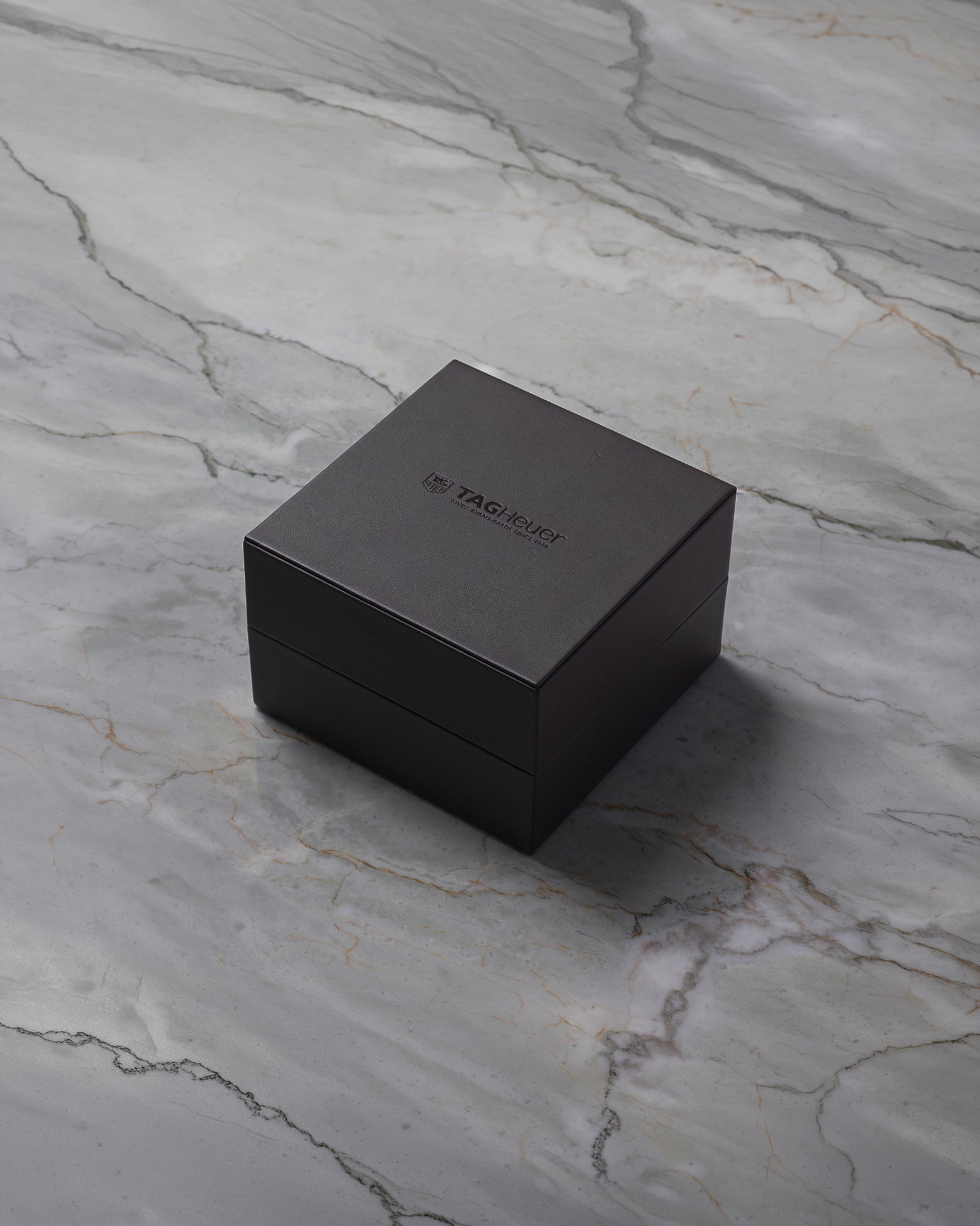 A closed square dark brown TAG Heuer luxury watch presentation box sitting centered on a textured grey marble surface