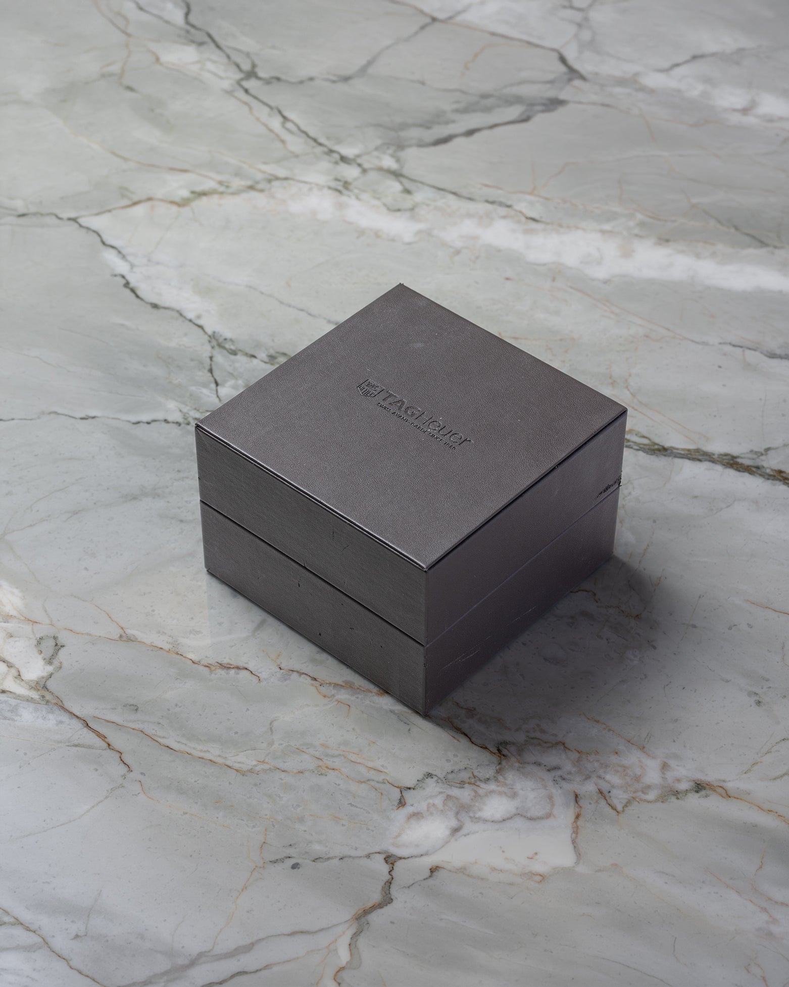 A closed square dark brown TAG Heuer luxury watch presentation box sitting on a textured grey marble background