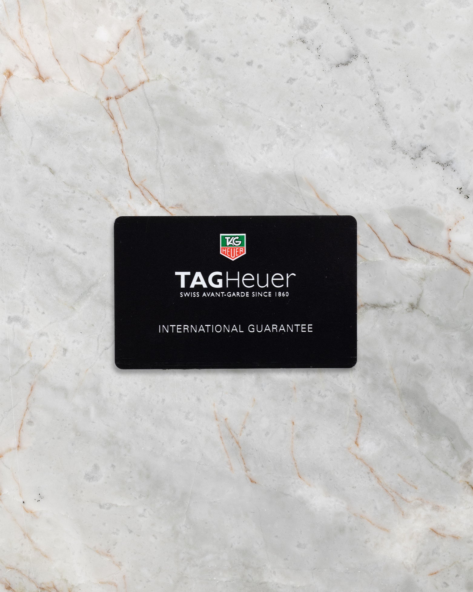 The official black TAG Heuer international guarantee warranty card displayed flat on a light marble background