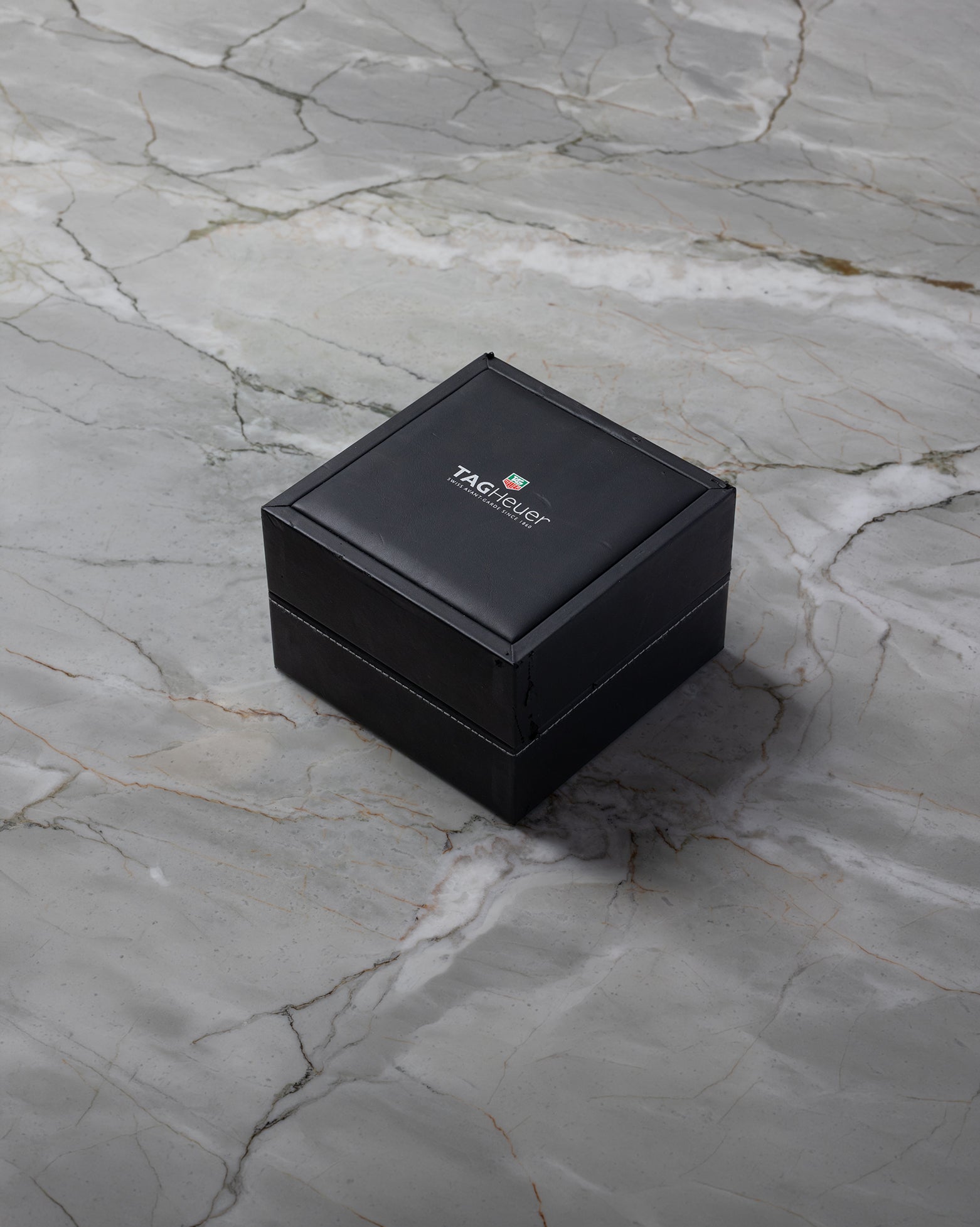 A closed square dark brown TAG Heuer luxury watch presentation box sitting centered on a textured grey marble background
