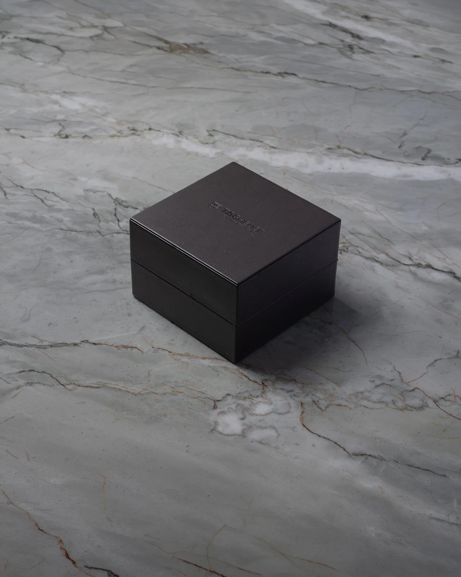 A top-down three-quarter angle of the official square black textured leatherette TAG Heuer watch presentation box resting on a grey marble backdrop
