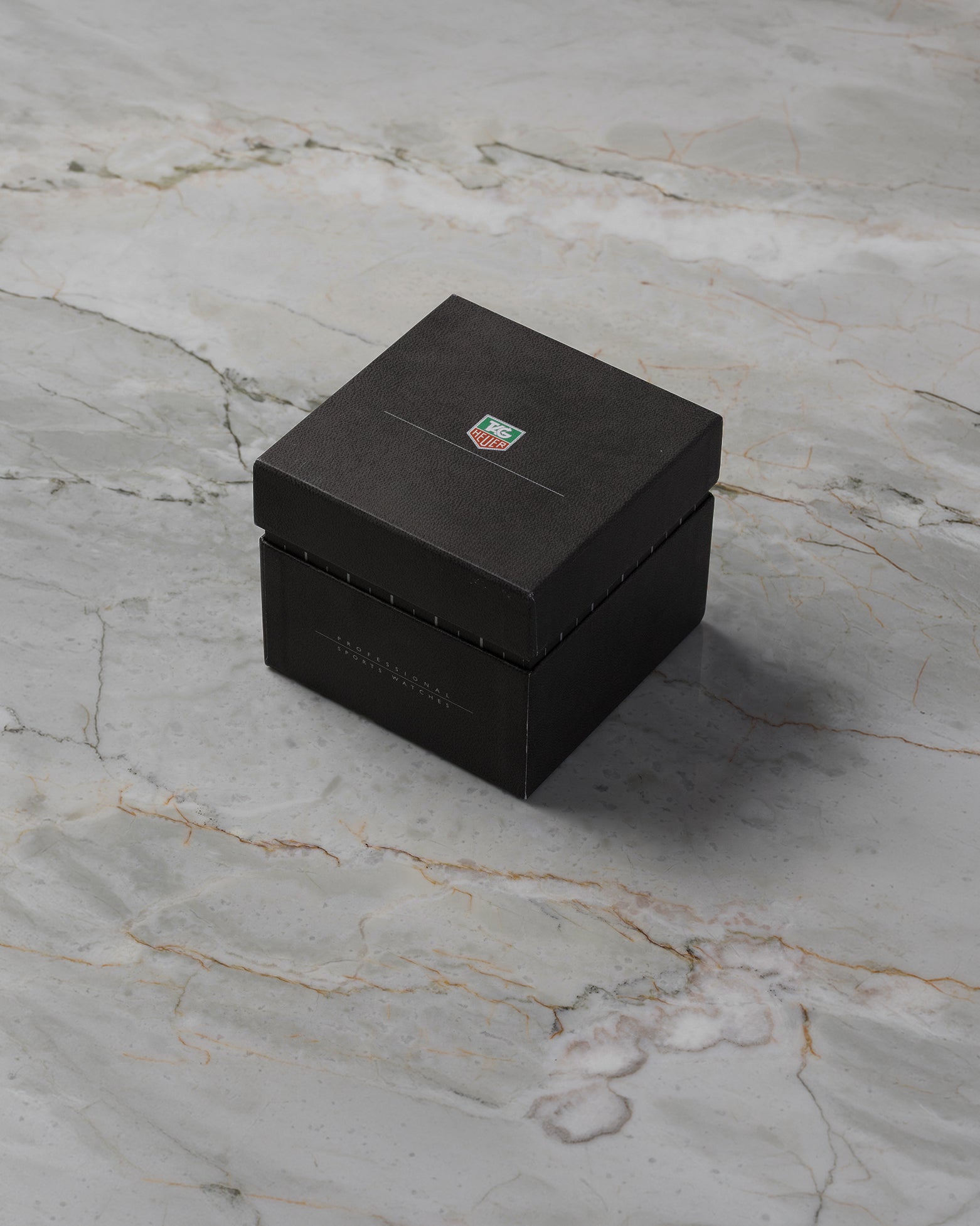 A high-angle three-quarter shot of the official vintage-style square black pebble-grain watch storage box featuring the colored brand shield crest on top