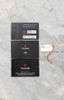 flat-lay view of the official Tudor warranty card, user instruction booklets, and white serial hangtag resting on light gray marble