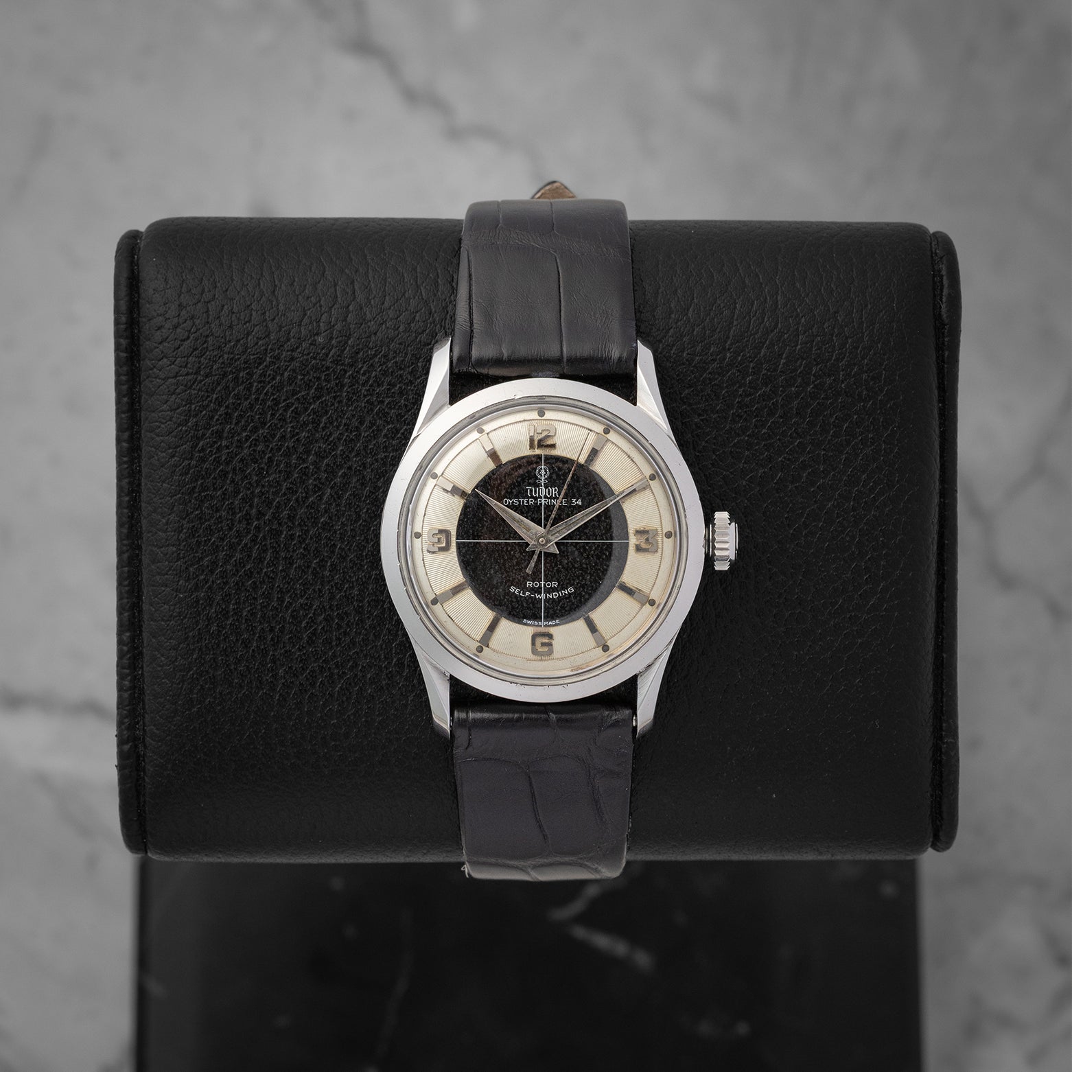 frontal portrait of a vintage 1950s Tudor Oyster Prince 7950 watch with a black and cream Tuxedo sector dial on a leather display mount